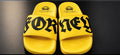 A pair of bright yellow slide sandals with bold black gothic-style lettering across the straps, displayed on a dark reflective surface. The city name 'FORNEY' is prominently printed on the straps, with 'INEFFABLE' and a crown logo visible on the footbeds.
