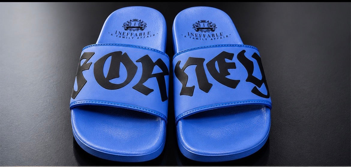 A pair of bright blue slide sandals with black Gothic-style lettering spelling 'FORNEY' across the straps and the Ineffable Logo printed on the heel.